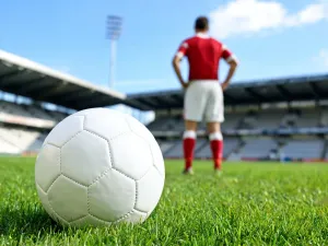 Bramkarz w kolorowym stroju stoi pewnie z piłką na murawie stadionu, na tle bramki i rozmytej publiczności.